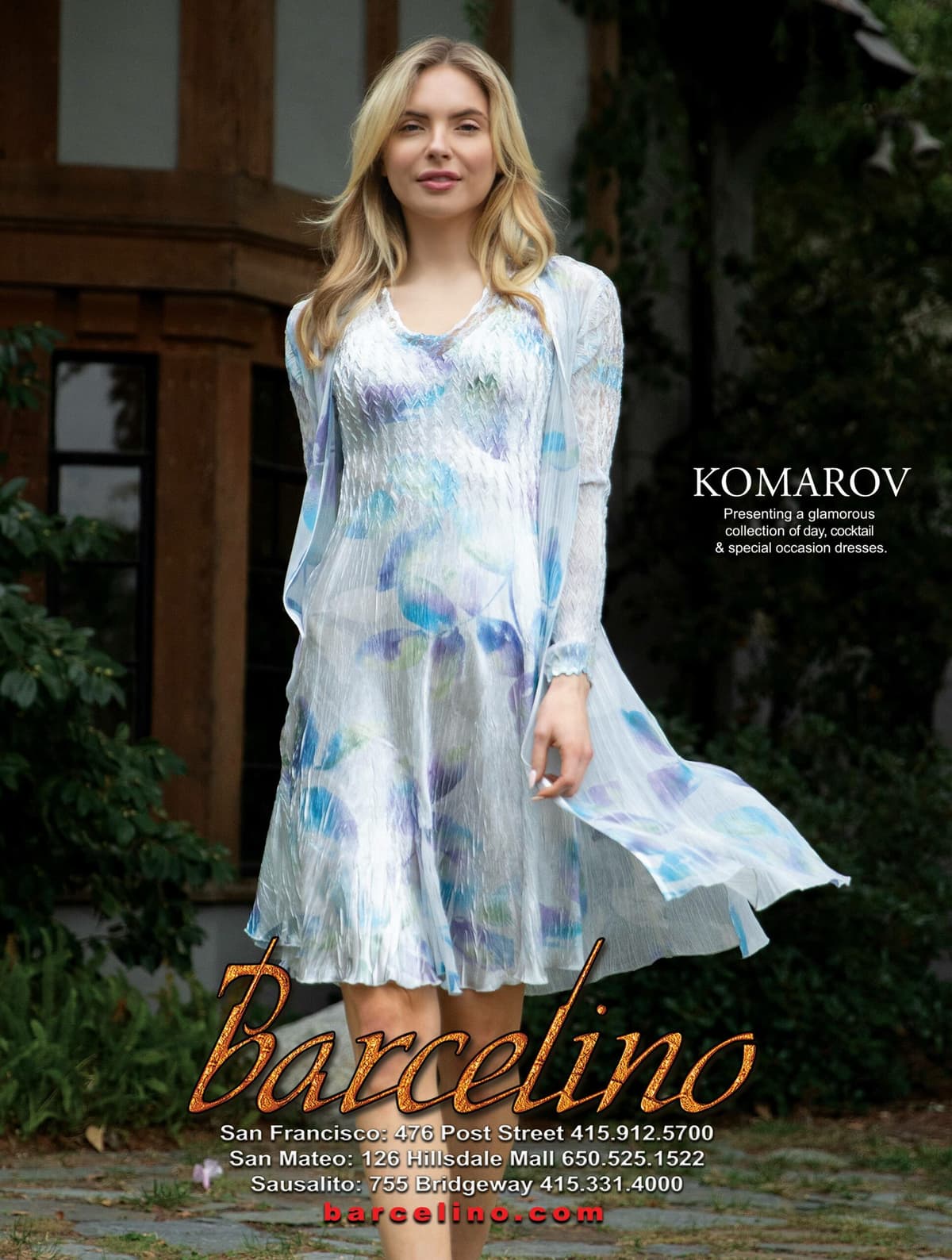 Elegant blonde woman modeling Barcelino's Komarov collection, wearing a flowing dress with blue and purple watercolor patterns, standing gracefully outdoors in front of a traditional building with ivy-covered walls, showcasing sophisticated fashion photography